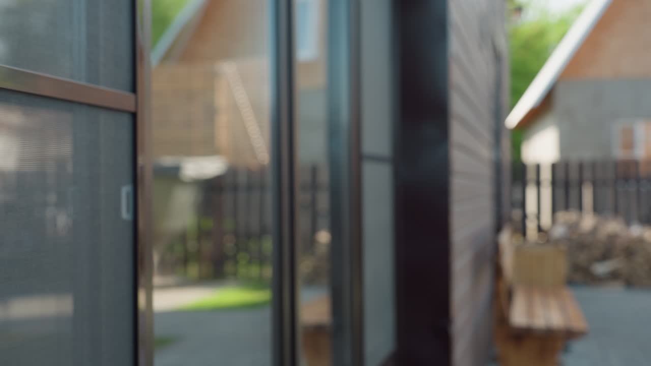 Close up of dark wood exterior siding with natural texture, reflective glass window showing faint outlines of opposite building, soft blur of outdoor bench, woodpile, and fence in sunlit background