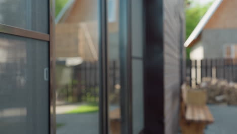 close up of dark wood exterior siding with natural texture, reflective glass window showing faint outlines of opposite building, soft blur of outdoor bench, woodpile, and fence in sunlit background