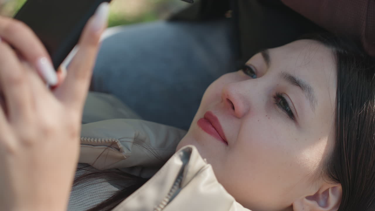 공원에서 스마트폰으로 메시지를 보며 미소짓는 아시아 여성, 편안한 디지털 대화와 몽상에 잠긴 듯한 표정을 클로즈업한 사진; 재킷에 부드러운 햇빛이 비치고 평화로운 여가 분위기가 느껴져요.