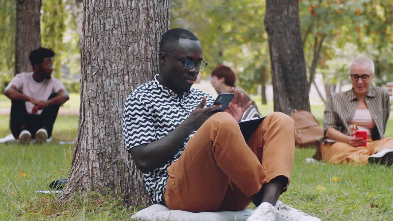 Young Afro Man Sitting in Park and Using Smartphone