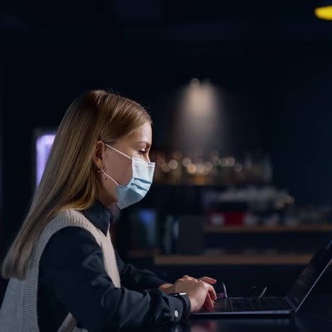 blonde woman wearing mask works at the computer in a dark room. lady working late in office. side view