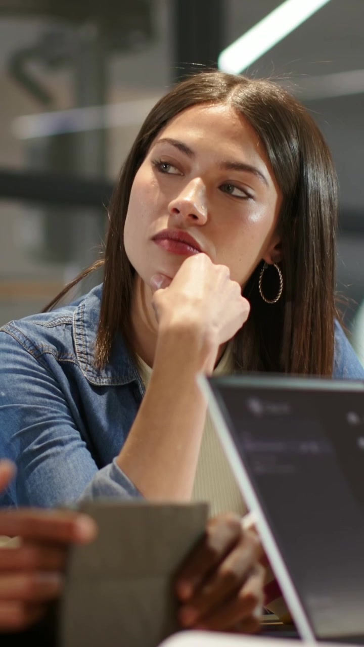 Video verticale: Donna in jeans che ascolta, osservando le mani di un collega indicare un tablet e un laptop, in fase di revisione.