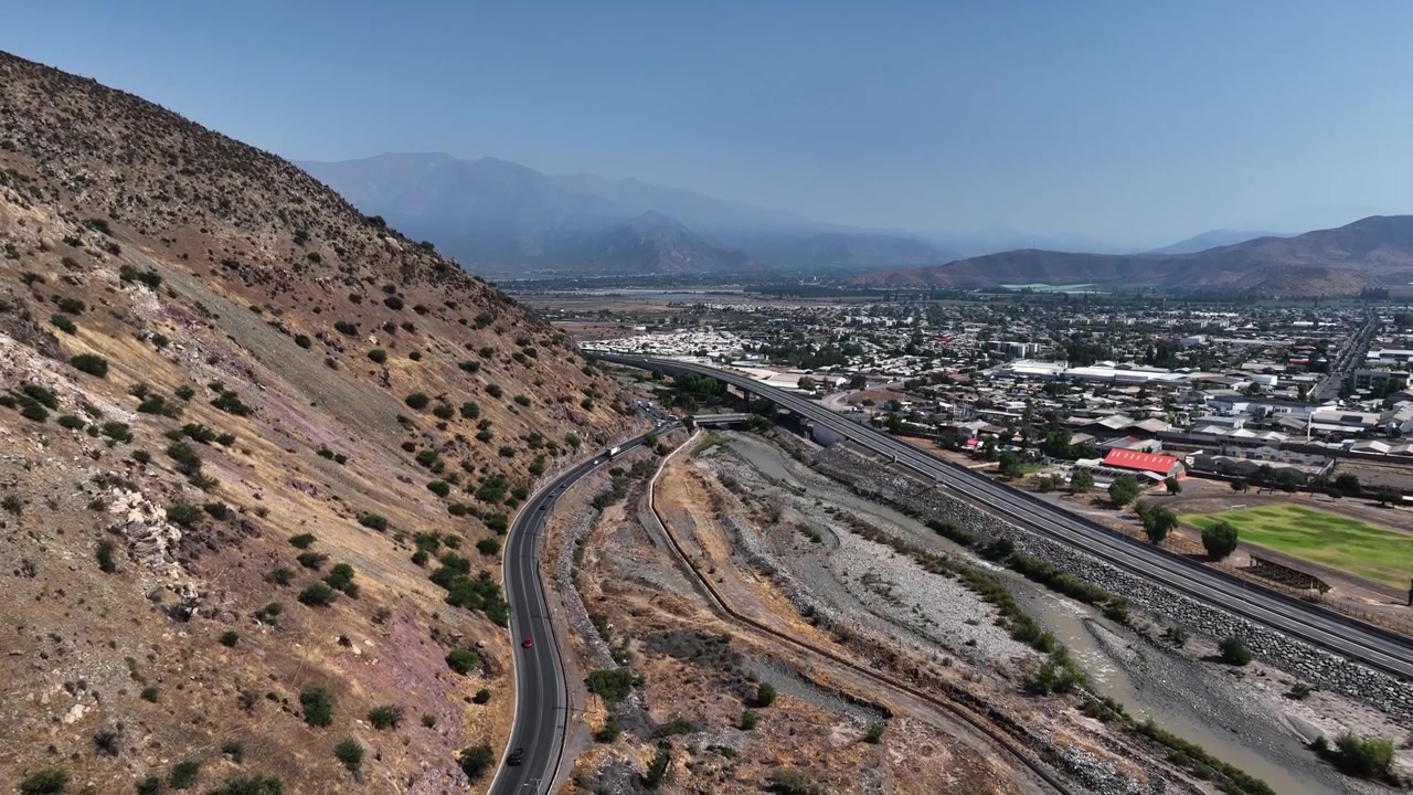 Ripresa aerea in discesa lungo un arido pendio montano che rivela un'autostrada tortuosa, un letto di fiume e l'intera città di Los Andes che si estende nella valle dell'Aconcagua, con le cime andine sullo sfondo. Los Andes, Cile.