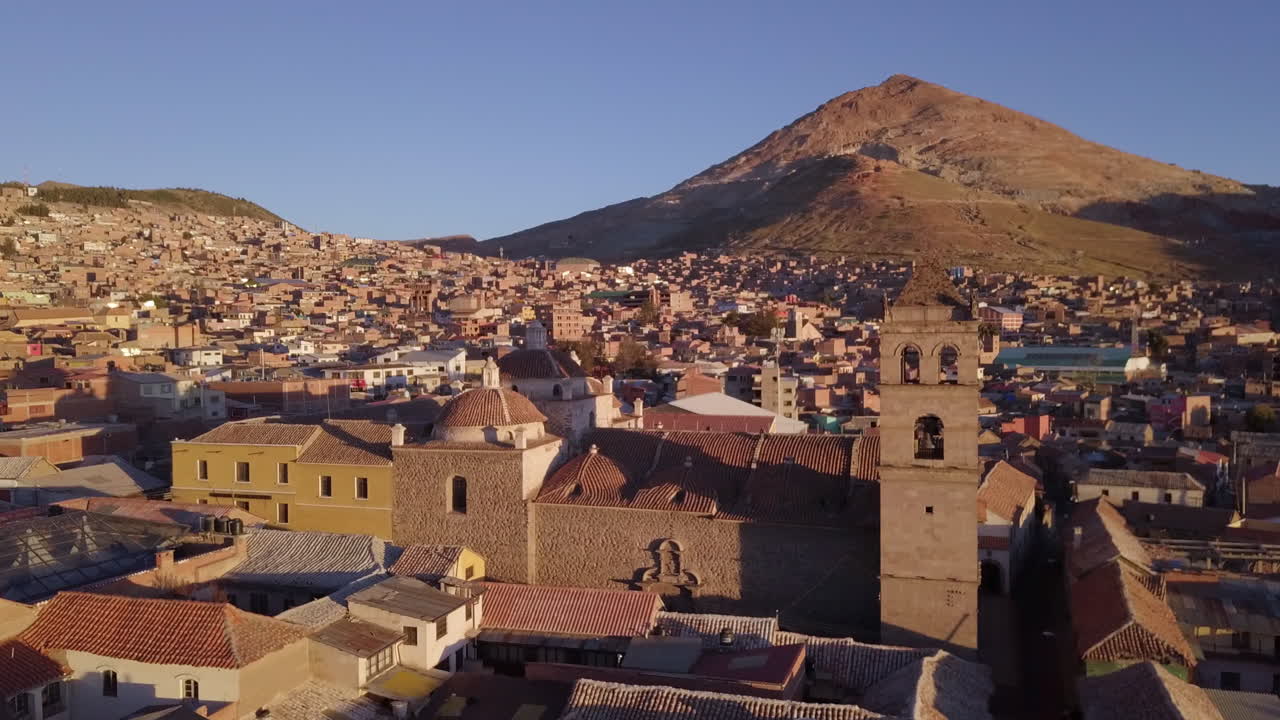 Ripresa con drone in volo in avanti sopra una città storica con architettura tradizionale e un campanile, con una grande montagna che si erge sullo sfondo.
