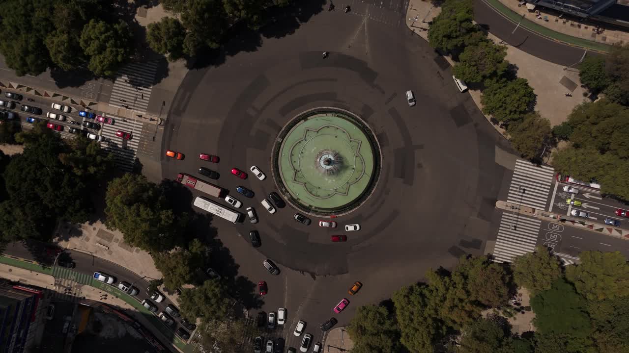 Top-down drone shot spinning above a busy roundabout fountain in Mexico City, showing traffic patterns and surrounding streets