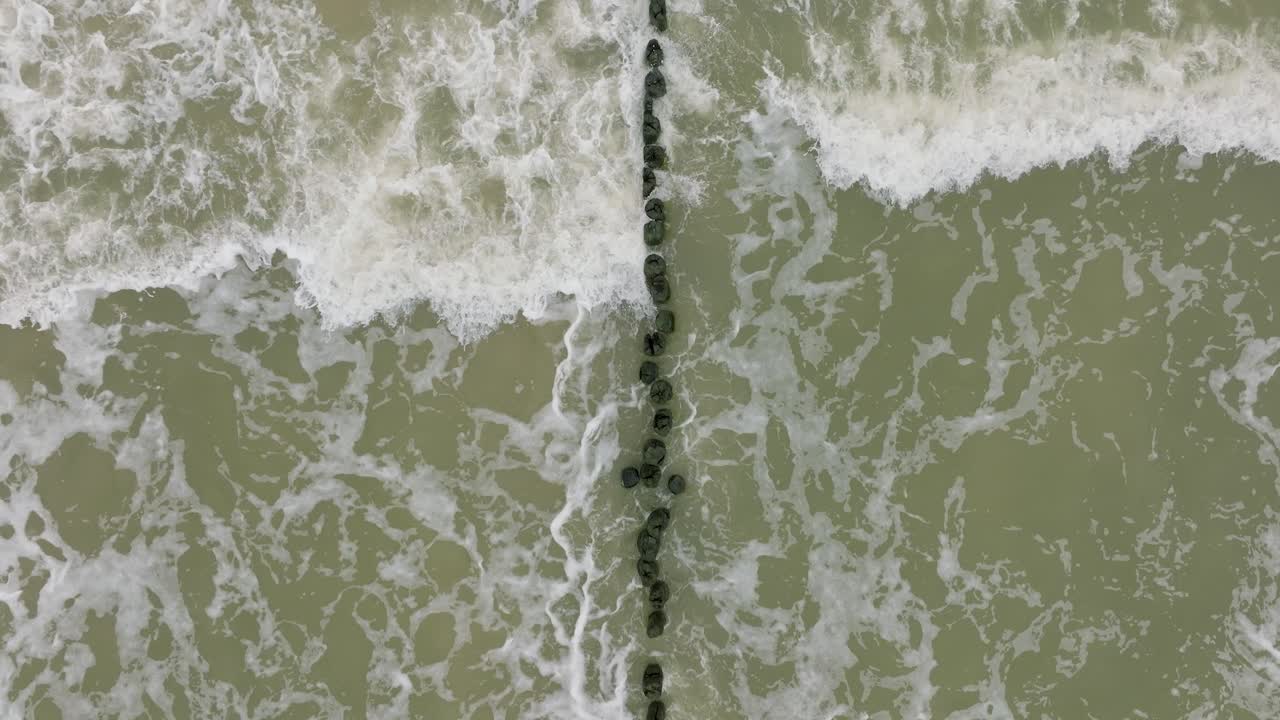 Aerial birdseye view of Baltic sea coast on a overcast day, old wooden pier, white sand beach, large storm waves crushing against the coast, climate changes, wide drone shot moving backward