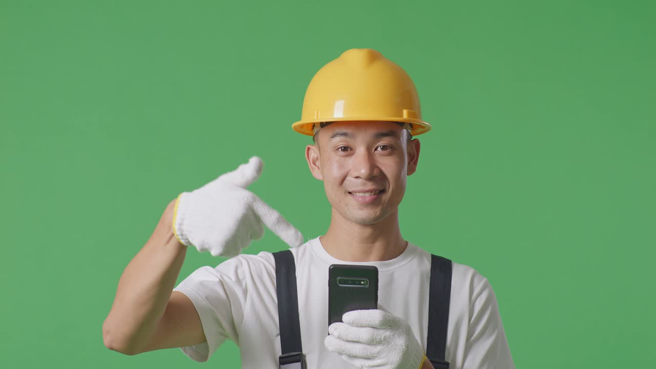 Close Up Of Asian Man Painter Wearing Safety Helmet Smiling And Pointing To Smartphone In His Hand While Standing In The Green Screen Background Studio
