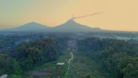 narrow river, awatu purbo waterfall and merapi volcano silhouette in background, aerial view