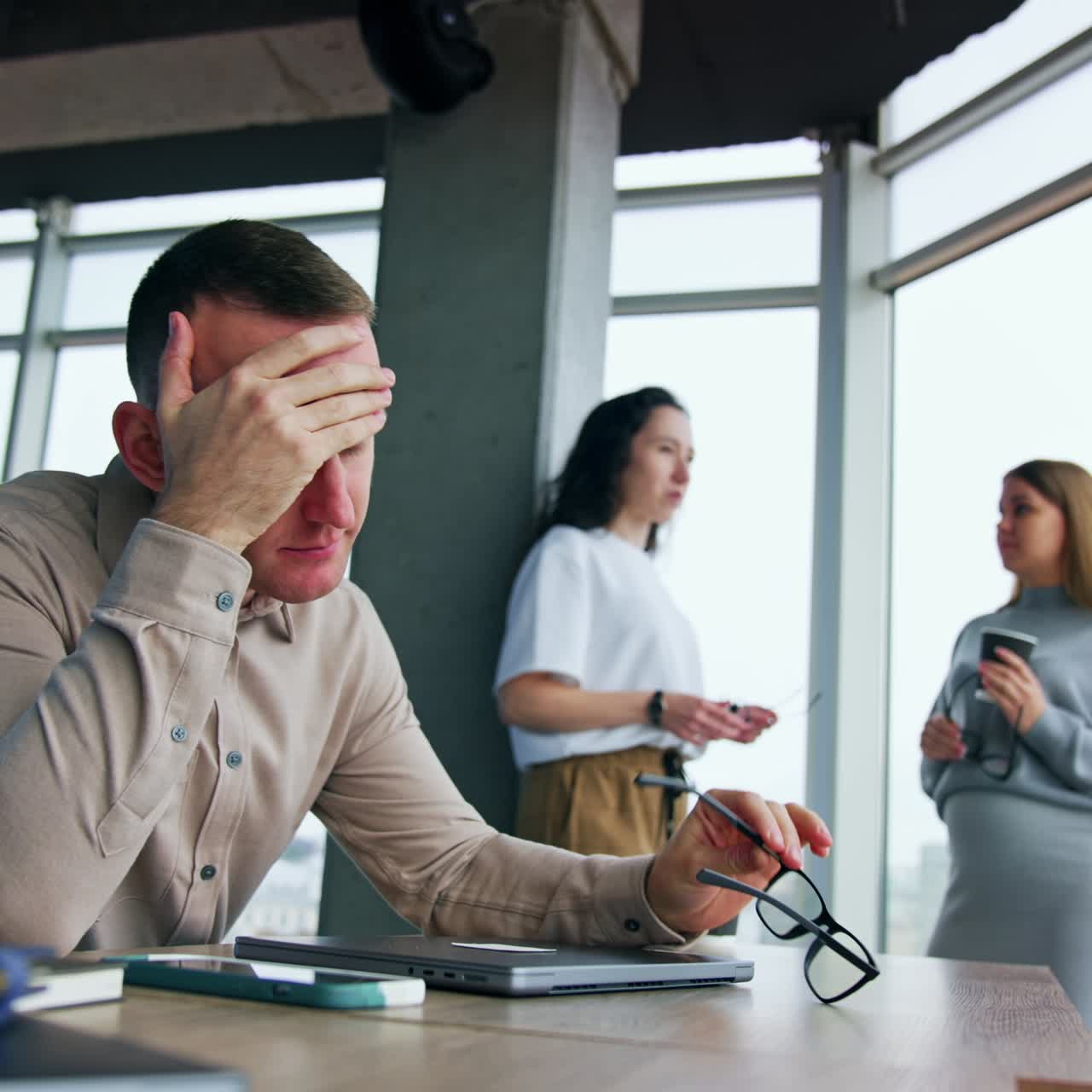Stressed tired male employee sits at desk. Man takes off his eyeglasses touching his head. Female colleagues talk at backdrop. Low angle view