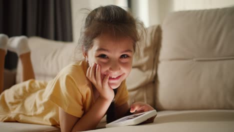 Close-up-shot:-Portrait-of-a-happy-little-brunette-girl-lying-on-a-light-brown-sofa-looking-at-something-on-her-phone-smiling-and-looking-at-the-camera-in-a-modern-room