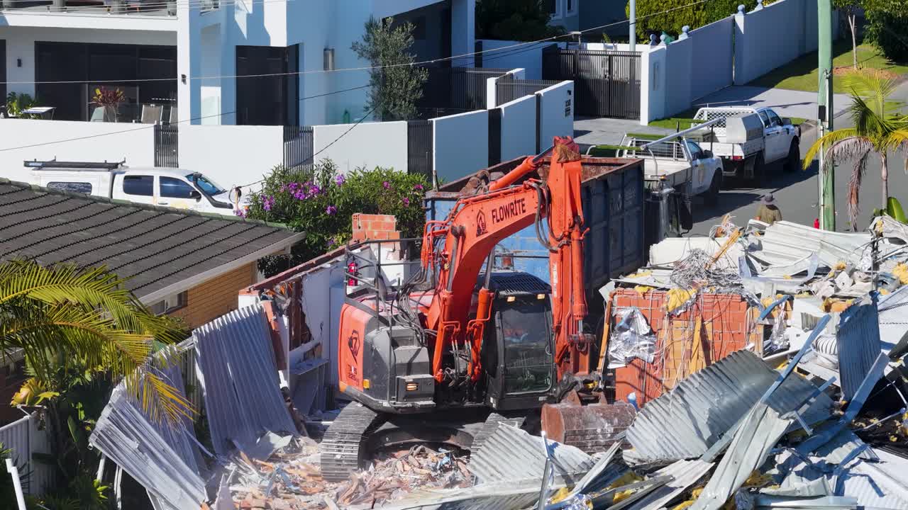 An orange excavator demolishes a residential house in daylight, surrounded by debris and construction workers, with modern homes and vehicles nearby. Static elevated camera