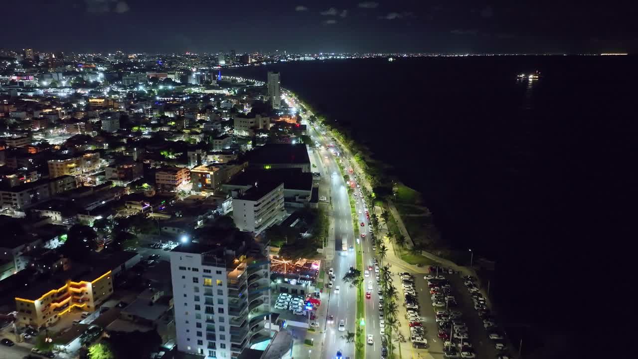 Cars on illuminated streets along coast of Santo Domingo city at night. Aerial Birds Eye shot. Illuminated city with apartment tower and houses in Dominican Republic. Wide shot. Peaceful scene