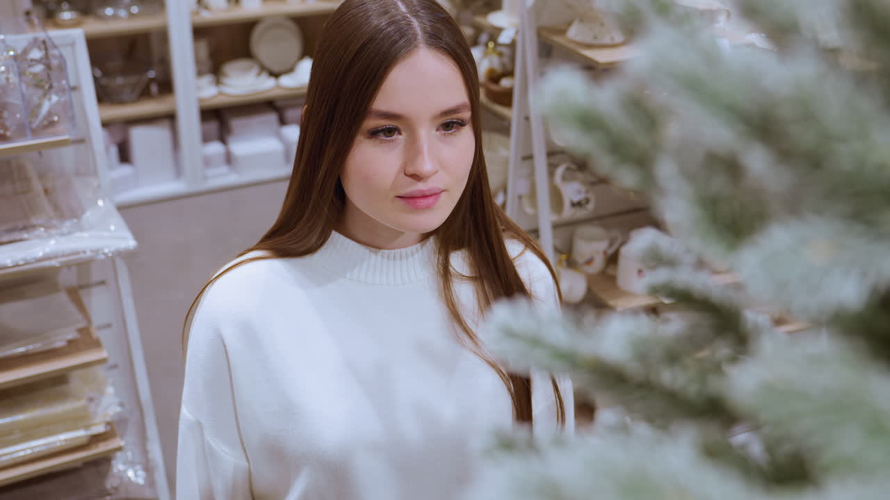 Lady standing in front of decorative plant, observing it thoughtfully with utensils displayed behind, creating a warm and peaceful shopping atmosphere in a well-lit home decor store