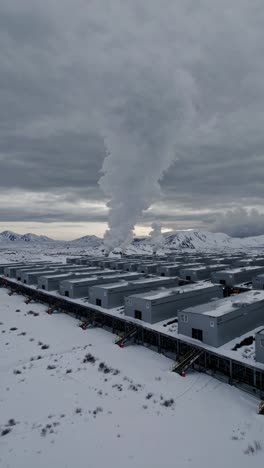 cryptocurrency mining farm revealing white vapor rising from identical gray buildings, set against snowy expanse and cloudy winter landscape with dramatic atmospheric perspective