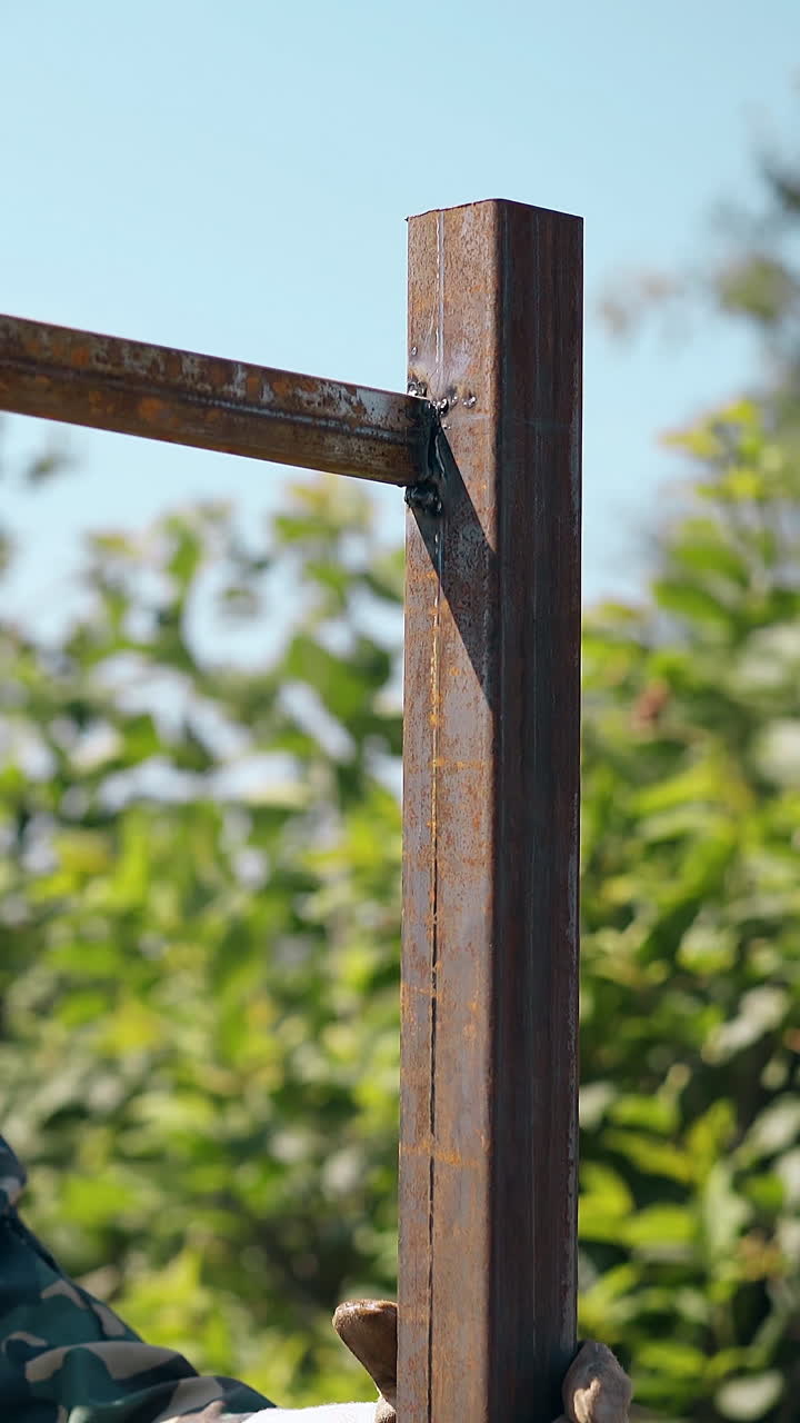 smiling worker in open welding mask taps on finished country house fence carcass with hammer on sunny day