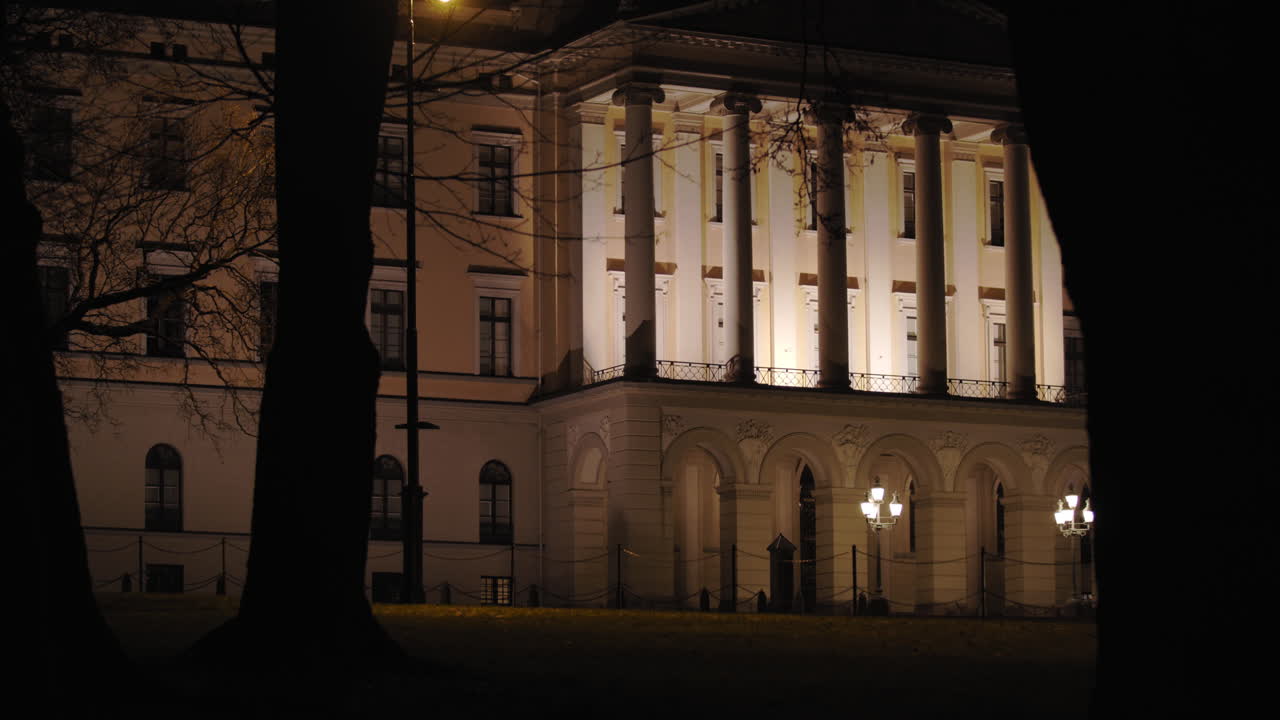Stabilized wide 4K shot of dark black trees moving from right to left in parallax motion in front of illuminated Norwegian Royal Palace building with iconic balcony, at night in Oslo Norway