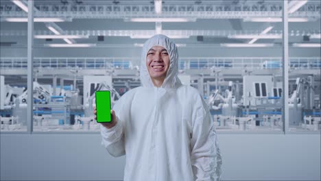 científico masculino asiático sonriendo y mostrando una pantalla verde de su teléfono inteligente mientras está de pie en una fábrica de laboratorio moderna con robots automatizados
