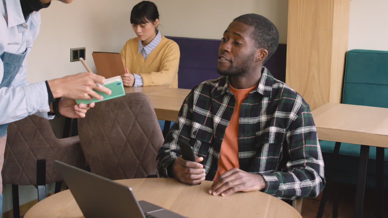 Happy Man Sitting At Table And Ordering Food To Waiter In Coffee Shop While Behind A Woman Working On Tablet