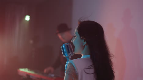 side view of a singer in a white gown singing and lifting her hands up, with a blurred keyboardist in the background. the scene is set against a soft red and blue background