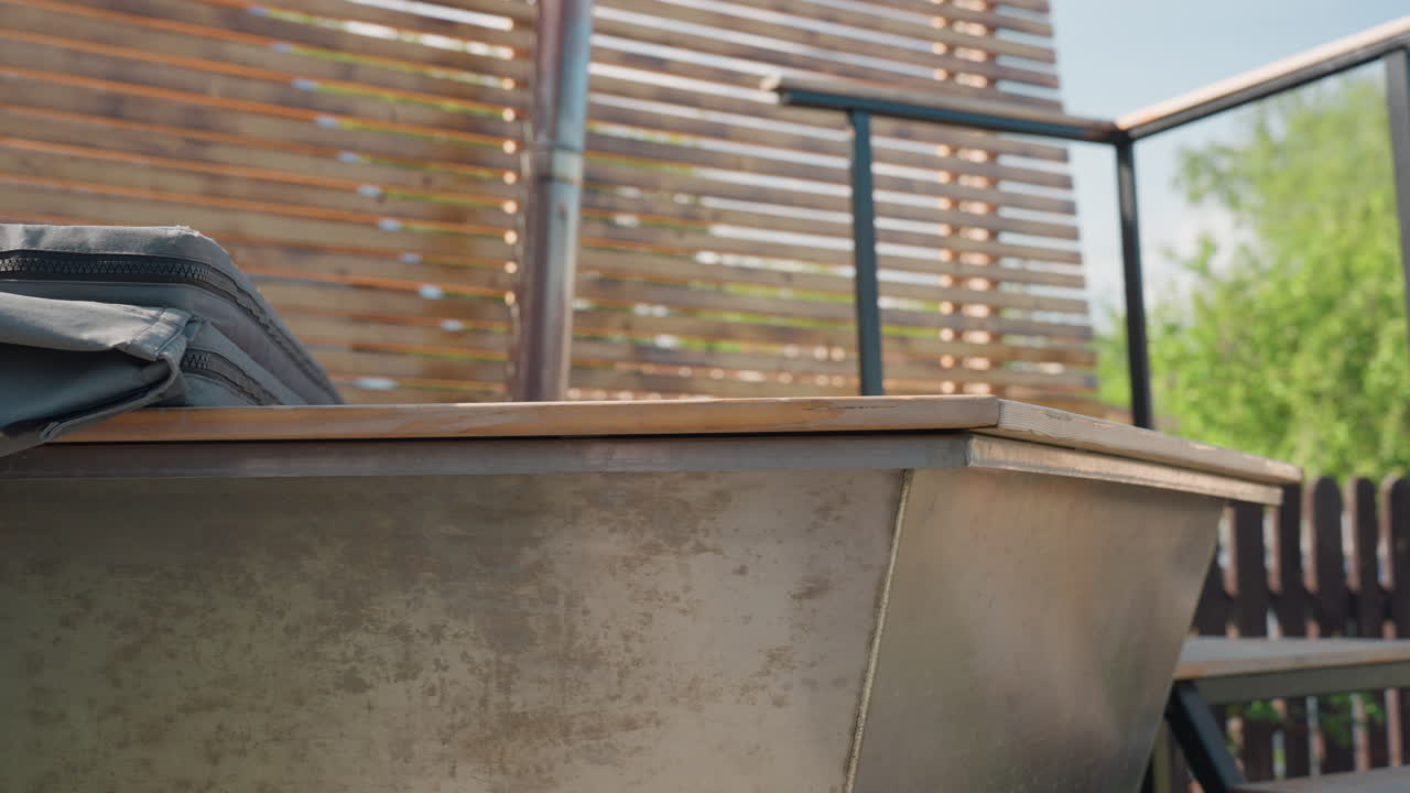 Close up of metal outdoor soaking tub with wooden rim and folded fabric cover placed beside slatted privacy fence, warm sunlight casting natural shadows across metallic surface and background