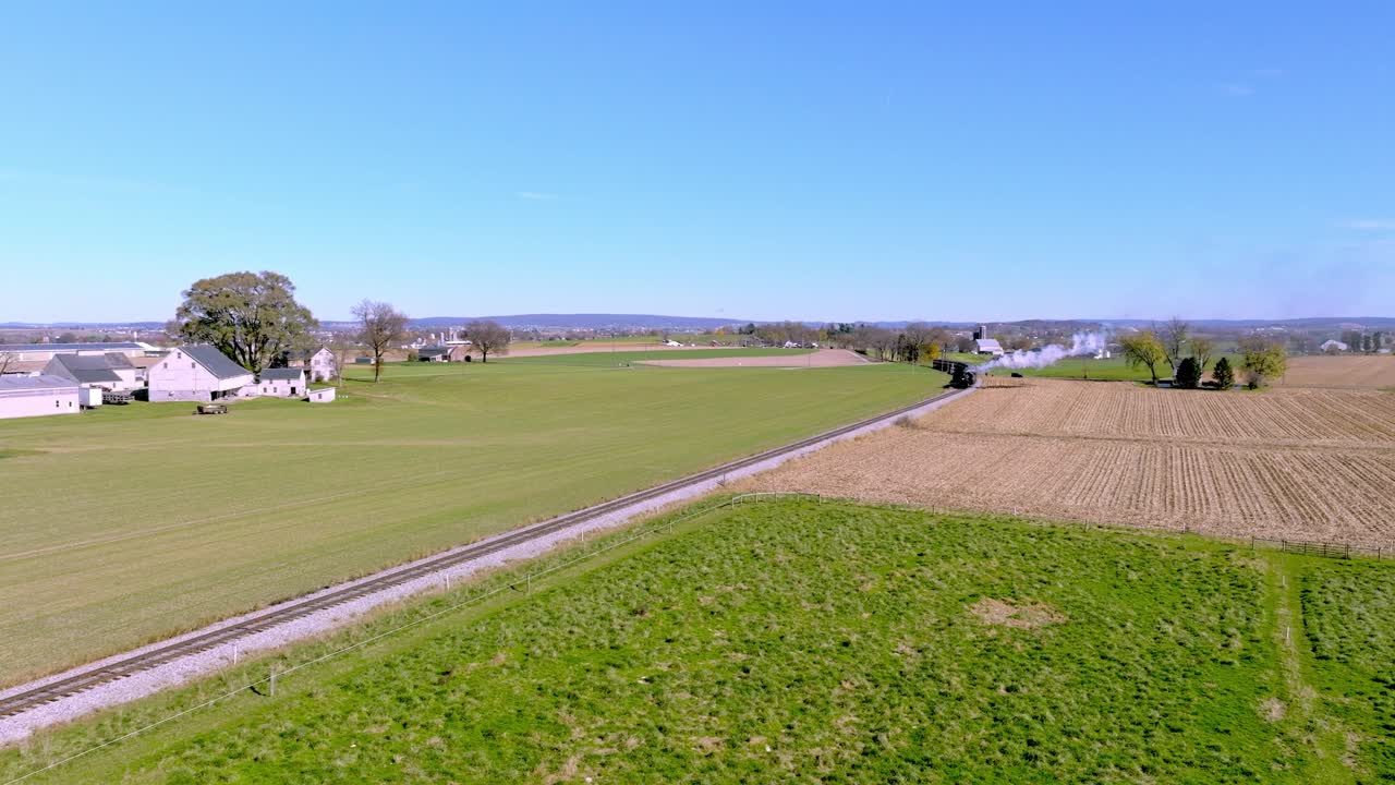 una vista aérea de una sola vía férrea que atraviesa tierras de cultivo mientras un tren de vapor se acerca en la distancia en un otoño soleado