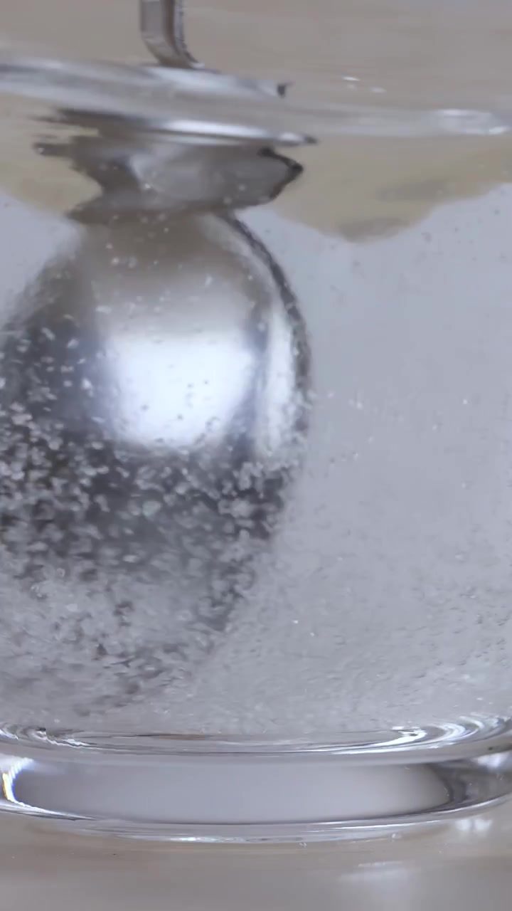 Primo piano di un cucchiaio di metallo che mescola lo zucchero nell'acqua in un bicchiere di vetro trasparente.