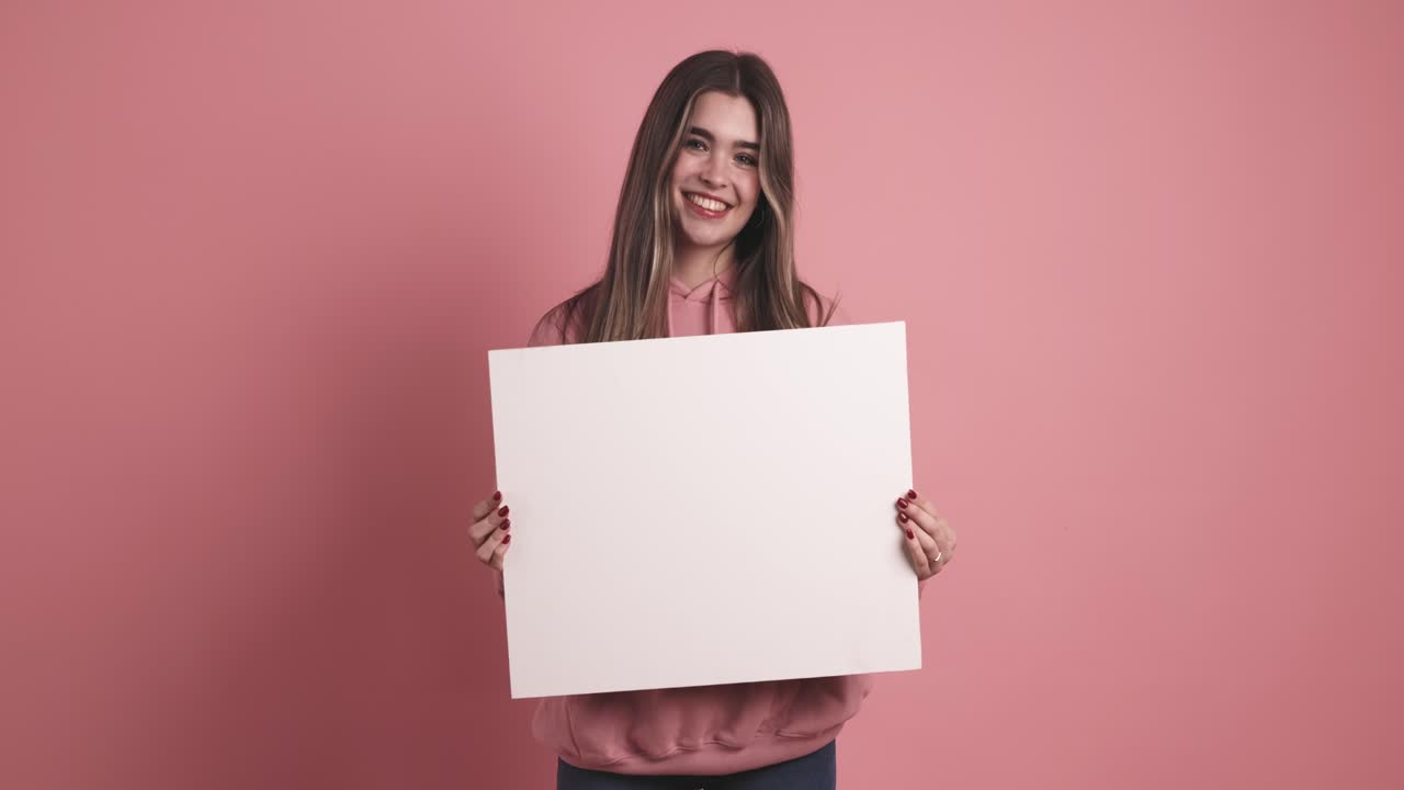 Happy young woman holding blank white paper in pink studio