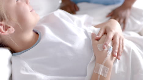 diverse female doctor and girl patient lying on bed holding hands in hospital, slow motion