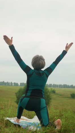 vista trasera de una mujer en traje verde y negro al aire libre en un vasto campo de hierba en una alfombra de yoga practicando una postura baja, brazos extendidos, bajo un cielo nublado con árboles alineados en el fondo
