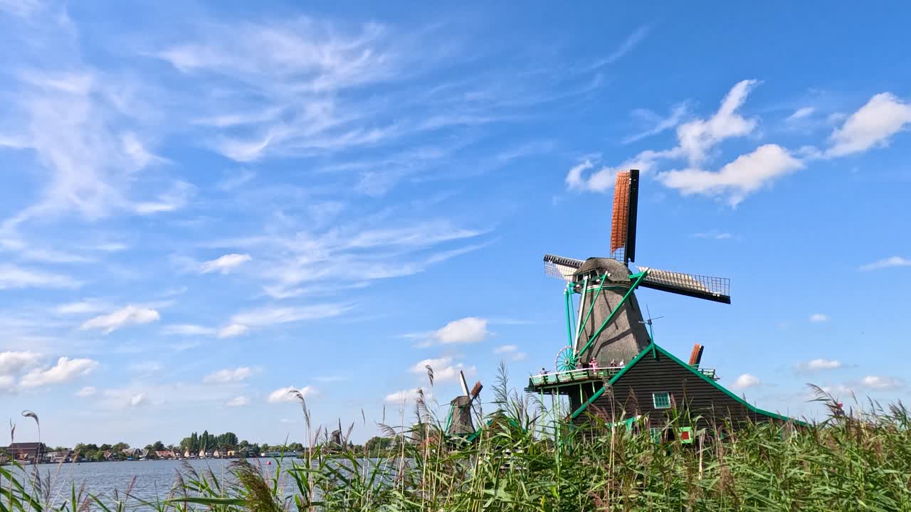 Historic windmill turns slowly by canal under bright blue sky, lush greenery, and natural daylight