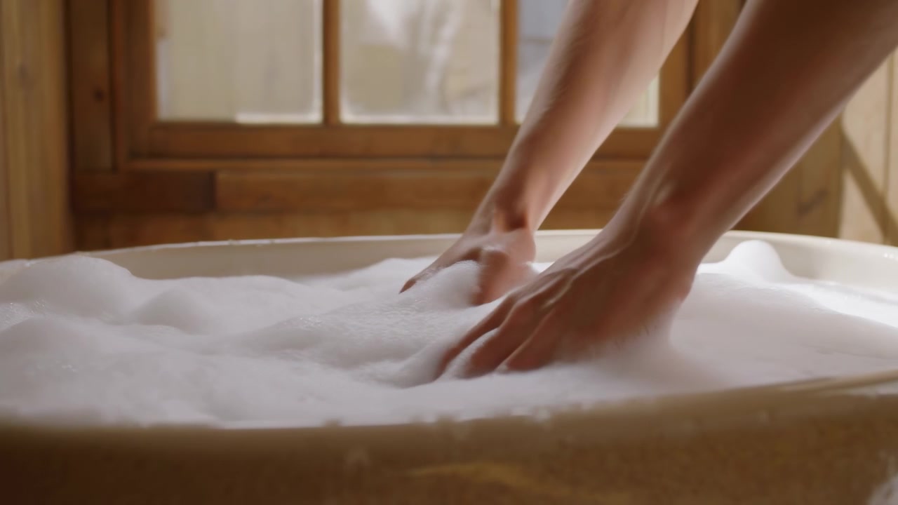 Close-up scene of hands and forearms pressing through dense white bath foam in rounded tub, creating ripples, displaced bubbles, soft reflections, and peaceful domestic spa atmosphere
