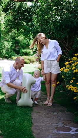 Long-haired Caucasian woman stands in the garden holding her little son by the hand. Happy dad runs up bringing a balloon of number one shape. Vertical video.
