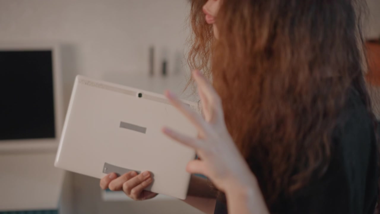 Profil d'une femme aux longs cheveux ondulés tenant un ordinateur portable blanc près de sa poitrine, se tournant dans un intérieur chaleureux. Un flou au niveau des doigts et l'inclinaison de l'appareil suggèrent un ajustement, une fermeture ou une présentation.