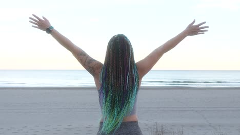 woman with braids breathes and spreads her arms on the beach at sunrise