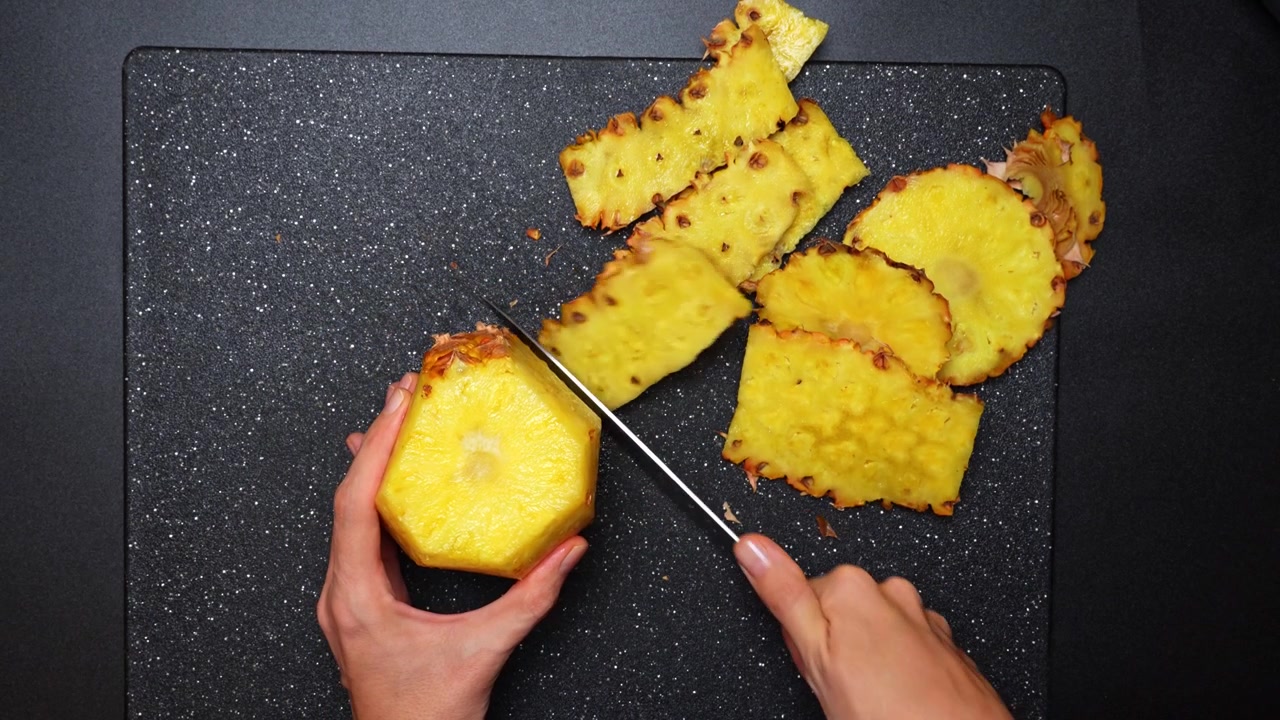 Person slicing fresh pineapple on a dark board. Top view of hands using a knife to cut the skin off a fresh yellow pineapple on a black surface.