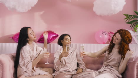 front view of three multiethnic friends toasting with champagne sitting on sofa celebrating bachelorette party