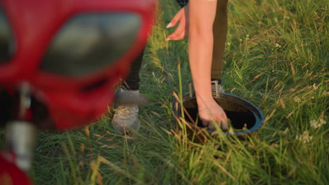 a close-up of a woman's hand squeezing a cloth, causing water to drip into a black bucket in a grassy field close to a bike