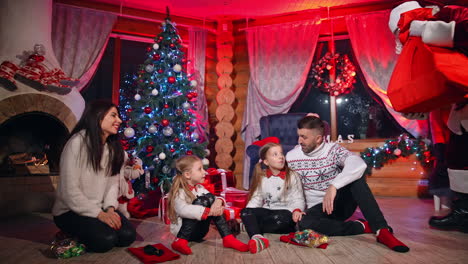cheerful family sitting near a fireplace and meeting santa with a big bag. kids rejoicing when seeing presents. santa comes to the family and sits beside them under the christmas tree.