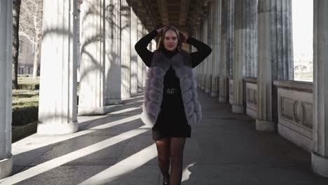 Blond-Girl-Walking-Through-Terrace-Corridor-Colonnade-Looking-At-Camera-and-Tidy-Hair-in-Berlin-City---gimbal-dolly-back-slow-motion