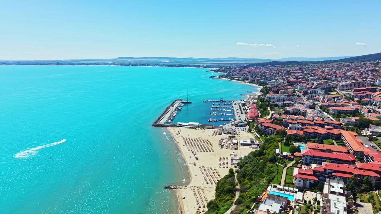 Güneşli bir günde, bir sahil kasabasında, kıyı boyunca şezlonglar, tekneler ve binalarla dolu bir plajın havadan görünümü.