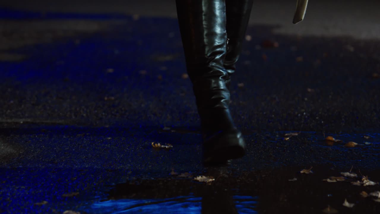 Primo piano degli stivali di una donna che calpestano una pozzanghera su una strada bagnata, riflettendo la luce neon blu di notte. Concetto per un'atmosfera urbana piovosa, uno stile da vita notturna e un'atmosfera cinematografica.