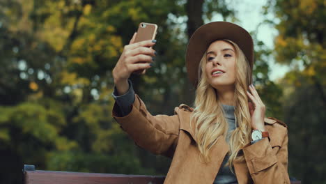 caucasian blonde caucasian woman sitting on the bench and taking a selfie with smartphone in the park in autumn