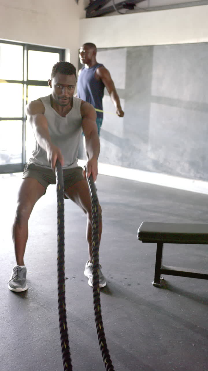 Vertical video: Men exercising with battle ropes and jump rope in gym, focused and determined