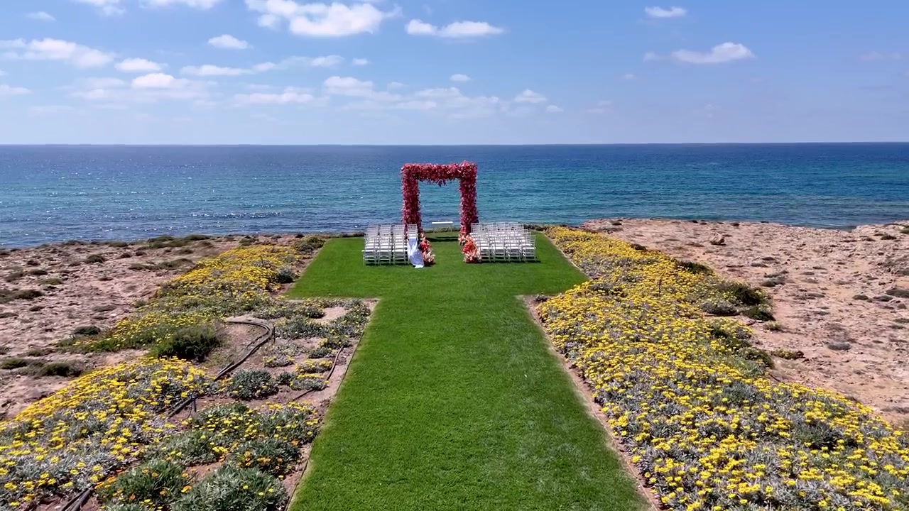 Un allestimento nuziale è organizzato vicino all'oceano, con file di sedie sull'erba verde. Fiori circondano l'area, creando uno spazio luminoso e invitante per un'occasione speciale
