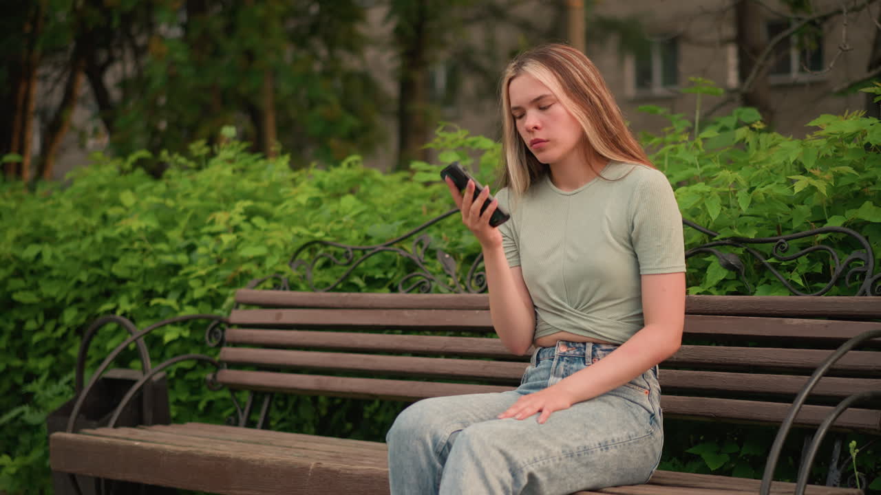 전화 통화 중 나무 벤치에 사려 깊게 앉아있는 젊은 여성, 왼손을 다리에 놓고 이마에 놓고, 울창한 초록색, 나무, 그리고 배경의 건물로 둘러싸여 있습니다.