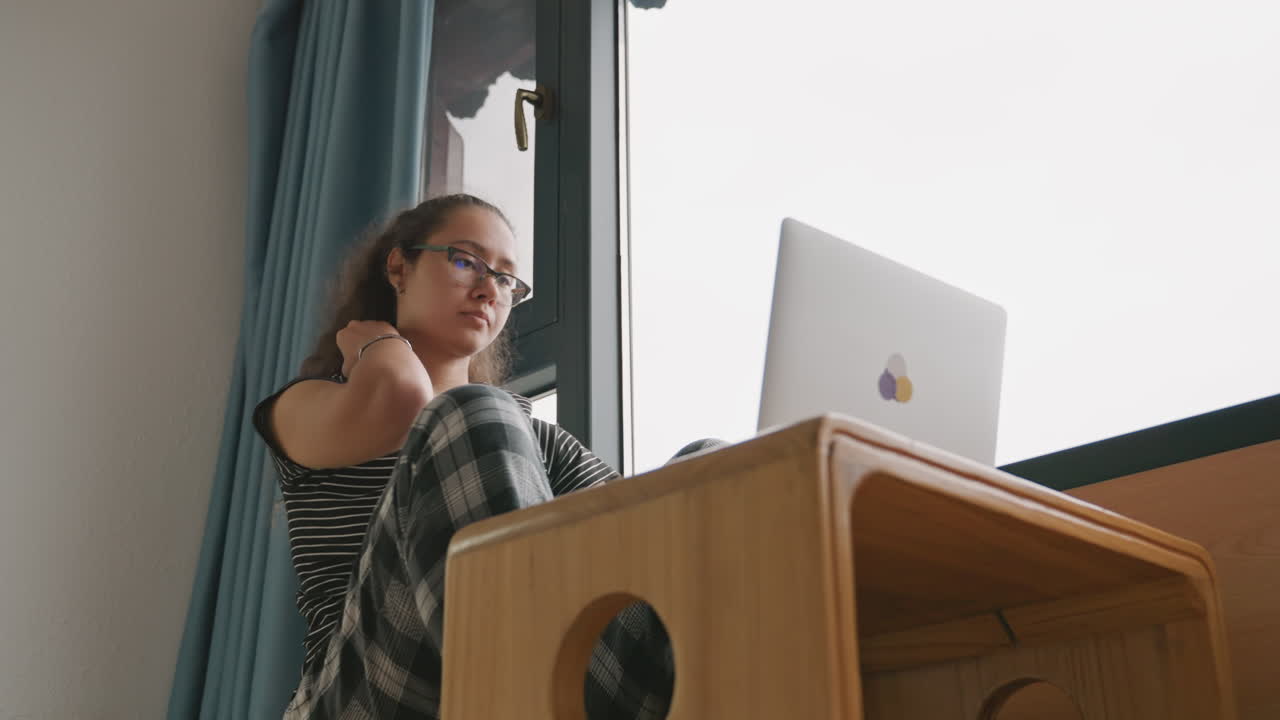 Scrivania di una camera d'albergo con laptop, riunione a distanza in corso. Una donna controlla la videocamera e si unisce a una teleconferenza. Scrivania in legno vicino alla finestra, postura professionale e rilassata, connessione stabile.