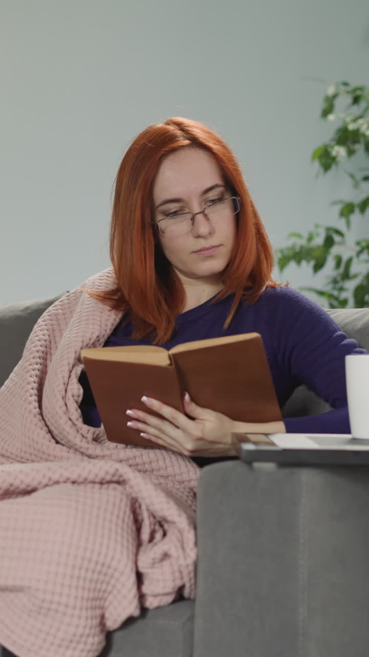 Redhead woman in glasses reads interesting book sitting on sofa in living room. Young housewife covered with blanket makes finger wet to flip pages