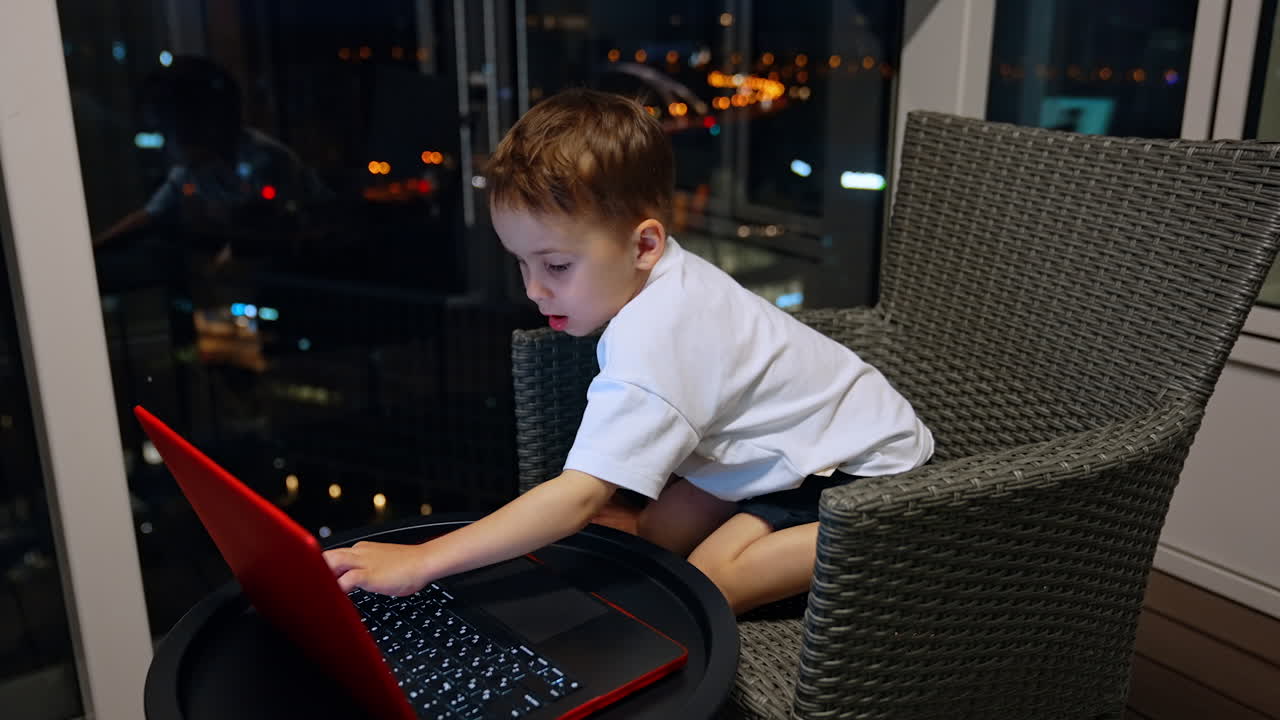 Child using laptop at night in city. A young boy sits on a chair, focused on typing on a laptop, with city lights glowing in the background at night