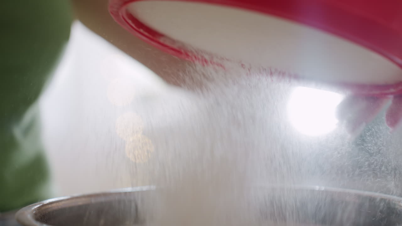 Close up of flour dust gently falling into stainless bowl during baking preparation, fine particles visible in soft light as cook works carefully to sift ingredients for homemade recipe