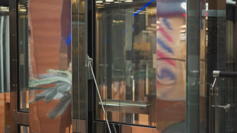 un moderno ascensor de vidrio que se mueve en un centro comercial, mostrando reflejos elegantes, detalles mecánicos y colores vibrantes borrosos de los interiores y anuncios circundantes